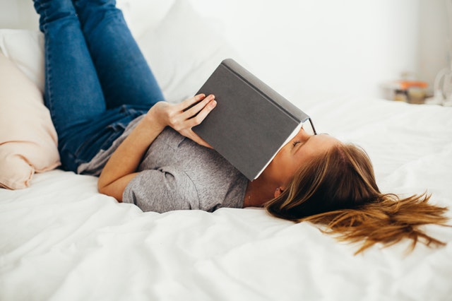 Žena leží so zdvihnutými nohami v posteli a na hlave má knihu.jpg