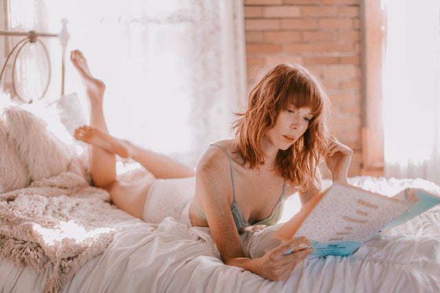 Ryšavá žena leží na posteli a číta časopis.jpg
